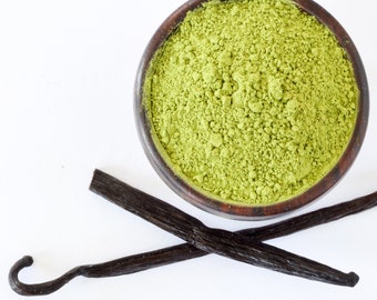 May include: A wooden bowl filled with green matcha powder, next to two vanilla beans on a white background.