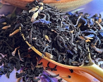 May include: Close-up of loose leaf tea with dried lavender buds and blue cornflower petals. The tea is dark brown and is in a gold spoon and a brown bowl. The background is a light purple color.
