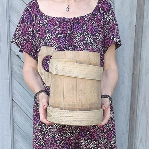 Puede incluir: Una taza de madera grande con asa, hecha de madera natural de color marrón claro. La taza está sostenida por una persona frente a una pared de madera.