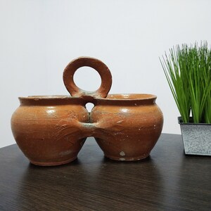 Puede incluir: Un juego de dos macetas de terracota con un asa que las conecta. Las macetas son de color marrón y tienen un aspecto rústico. Las macetas están sobre una superficie de madera marrón oscuro. Una pequeña planta en maceta está en el fondo.