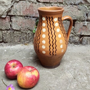 May include: A brown ceramic pitcher with a handle, decorated with white dots and black wavy lines. Two red apples and a purple flower are in the foreground. The pitcher is on a concrete surface with a brick wall in the background.