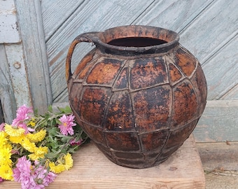 May include: A rustic brown ceramic pitcher with a textured, woven design sits on a wooden stool. A bouquet of yellow and pink flowers is in front of the pitcher.