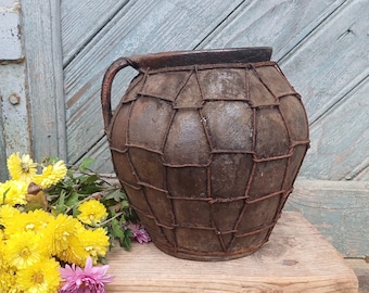 May include: A rustic brown clay pot with a woven wire design sits on a wooden shelf with a bouquet of yellow and pink flowers.