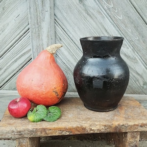 May include: A black ceramic jug and a bright orange gourd on a wooden table. The jug has a rounded body and a wide mouth. The gourd has a smooth, slightly bumpy surface.