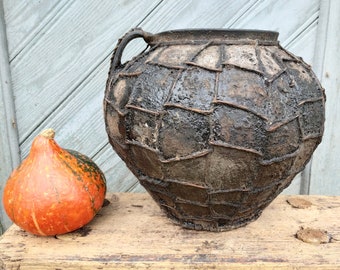 May include: A large, dark brown, ceramic pot with a woven pattern and a handle on each side. The pot is sitting on a wooden stool next to a small orange pumpkin.