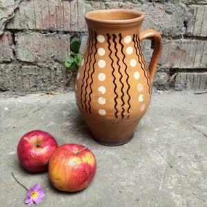 May include: A brown ceramic pitcher with a handle, decorated with white dots and black wavy lines. Two red apples with yellow accents and a small purple flower rest on a concrete surface near the pitcher.