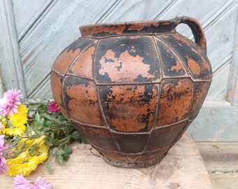 May include: A large, brown clay pot with a handle. The pot is decorated with a black grid pattern and has a rustic, aged appearance.