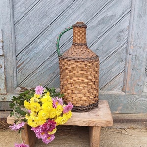 May include: A woven wicker bottle with a green and red handle sits on a wooden stool. A bouquet of yellow and pink chrysanthemums sits in front of the bottle.