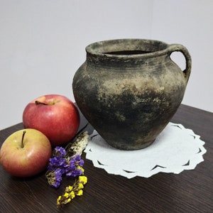 Può includere: Un vaso di terracotta marrone rustico con manico su un tovagliolo bianco con due mele rosse e fiori viola e gialli.
