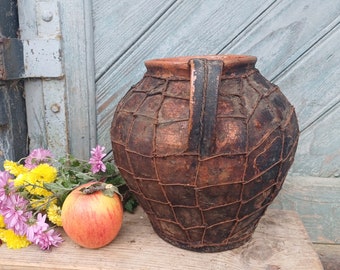 May include: A rustic brown clay pot with a woven design and a handle. The pot is sitting on a wooden surface with a bouquet of yellow and pink flowers and a red apple.