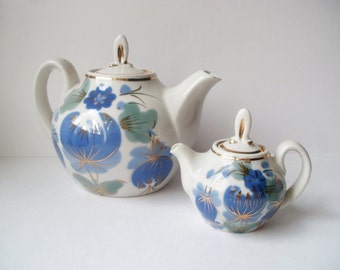 Puede incluir: Un juego de dos teteras de porcelana blanca con diseños florales azules y dorados. La tetera más grande tiene un cuerpo redondo y un pico en el lateral. La tetera más pequeña es una versión en miniatura de la más grande.