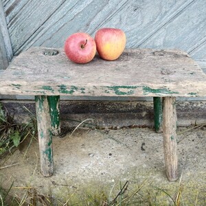 Puede incluir: Un taburete de madera r&uacute;stico con detalles de pintura verde y dos manzanas rojas en la parte superior. El taburete tiene un aspecto desgastado y est&aacute; sobre una superficie de hormig&oacute;n.