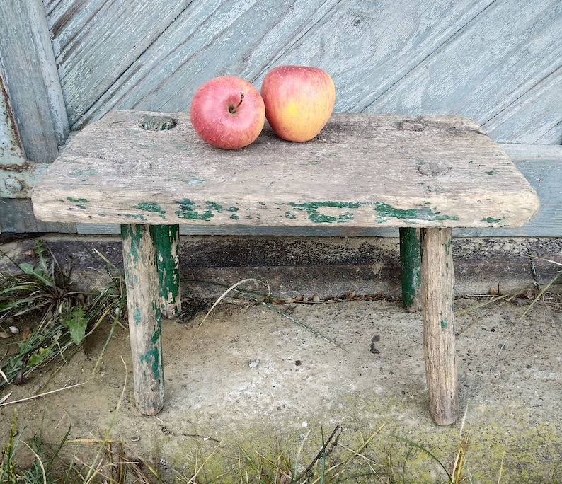 Puede incluir: Un taburete de madera r&uacute;stico con detalles de pintura verde y dos manzanas rojas en la parte superior. El taburete tiene un aspecto desgastado y est&aacute; sobre una superficie de hormig&oacute;n.