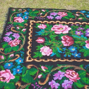 May include: A black wool rug with a floral pattern. The rug features pink, red, blue, and purple flowers on a black background. The rug has a gold border.