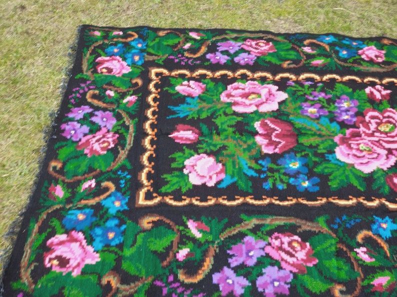 May include: A black wool rug with a floral pattern. The rug features pink, red, blue, and purple flowers on a black background. The rug has a gold border.