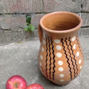 May include: A handmade, terracotta-colored pitcher with a handle, decorated with white polka dots and black wavy lines. Two red apples rest near the base of the pitcher. The pitcher is likely used for serving beverages.