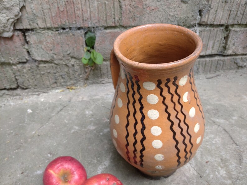 May include: A handmade, terracotta-colored pitcher with a handle, decorated with white polka dots and black wavy lines. Two red apples rest near the base of the pitcher. The pitcher is likely used for serving beverages.