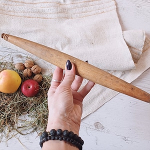 May include: A wooden rolling pin with a natural finish. The rolling pin is approximately 45 cm long and has a slightly rounded handle. The rolling pin is lying on a white surface with a white cloth and some dried hay.