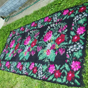 May include: A black rug with a floral pattern in shades of pink, red, and green. The rug has a fringe border.