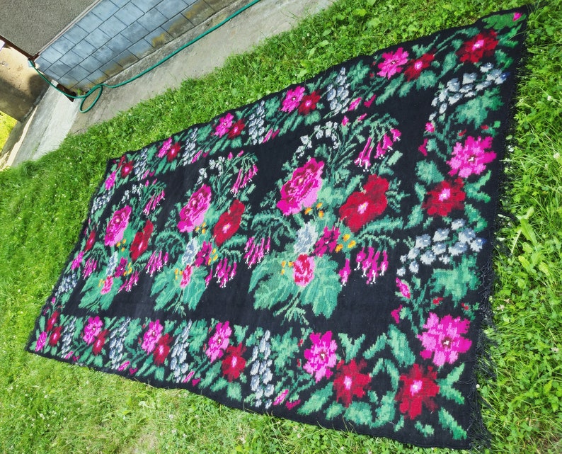 May include: A black rug with a floral pattern in shades of pink, red, and green. The rug has a fringe border.
