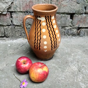 May include: A brown ceramic pitcher with a handle, decorated with black wavy lines and white dots. Two red apples with yellow streaks and a small purple flower are in front of the pitcher. The pitcher is likely used for serving water or beverages.