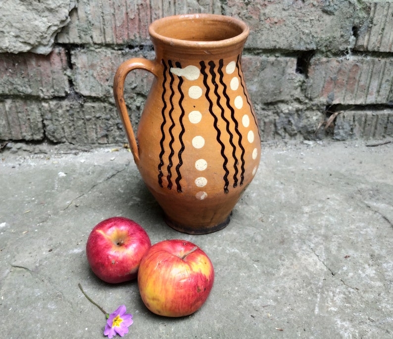 May include: A brown ceramic pitcher with a handle, decorated with black wavy lines and white dots. Two red apples with yellow streaks and a small purple flower are in front of the pitcher. The pitcher is likely used for serving water or beverages.