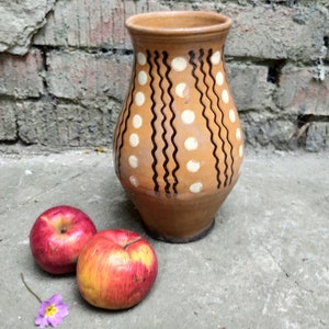 May include: A brown ceramic vase with a wide base and a narrow neck, decorated with white polka dots and black wavy lines. Two red apples and a small purple flower are in front of the vase, set against a weathered brick wall.