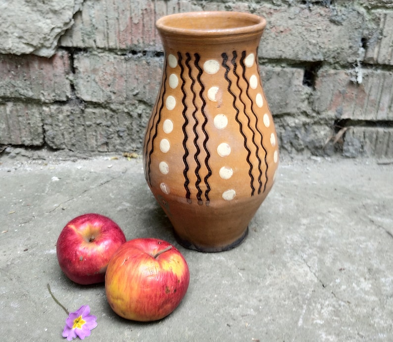 May include: A brown ceramic vase with a wide base and a narrow neck, decorated with white polka dots and black wavy lines. Two red apples and a small purple flower are in front of the vase, set against a weathered brick wall.