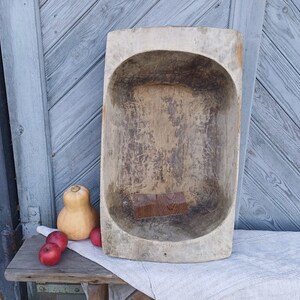 May include: A large, wooden, oval-shaped dough bowl with a worn, rustic finish. The bowl is light brown in color and has a natural wood grain. It is sitting on a white cloth with three red apples and a small, orange gourd.