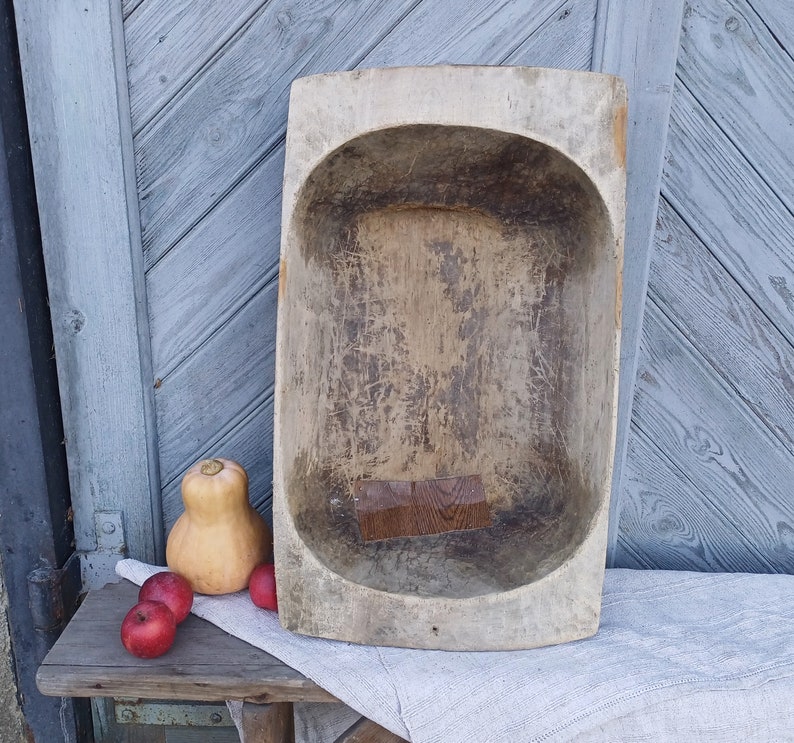 May include: A large, wooden, oval-shaped dough bowl with a worn, rustic finish. The bowl is light brown in color and has a natural wood grain. It is sitting on a white cloth with three red apples and a small, orange gourd.