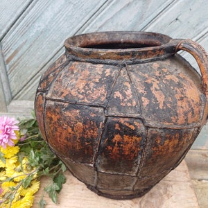 May include: A large, rustic, brown clay pot with a wide opening and a single handle. The pot has a textured, cracked surface and appears to be very old.