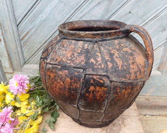 May include: A large, rustic, brown clay pot with a wide opening and a single handle. The pot has a textured, cracked surface and appears to be very old.