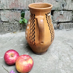 May include: A handmade, tan-colored ceramic pitcher with a handle, decorated with black wavy lines and white dots. Two red apples with yellow accents rest on a concrete surface near the pitcher. The background features a weathered brick wall.