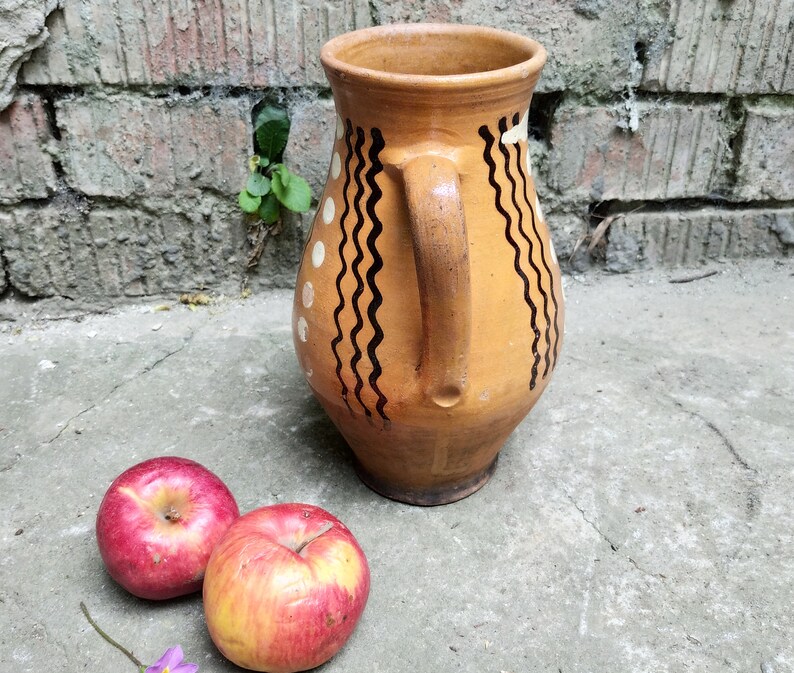 May include: A handmade, tan-colored ceramic pitcher with a handle, decorated with black wavy lines and white dots. Two red apples with yellow accents rest on a concrete surface near the pitcher. The background features a weathered brick wall.