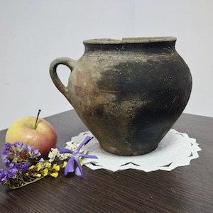 May include: A rustic, dark brown clay pot with a handle. The pot is sitting on a white doily on a dark wood surface. There are purple and yellow flowers and a red apple in the foreground.