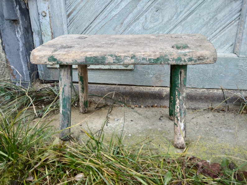 Puede incluir: Un taburete de madera r&uacute;stico con tres patas pintadas de verde. El taburete est&aacute; desgastado y muestra signos de antig&uuml;edad. Est&aacute; sentado sobre una superficie de hormig&oacute;n con hierba en primer plano.