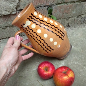 May include: A rustic, hand-painted ceramic pitcher with a brown base color, adorned with white polka dots and black wavy lines. The pitcher has a handle and is accompanied by two red apples. The pitcher is likely used for serving beverages.