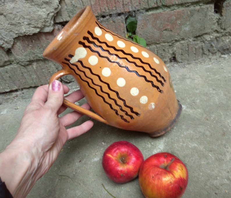 May include: A rustic, hand-painted ceramic pitcher with a brown base color, adorned with white polka dots and black wavy lines. The pitcher has a handle and is accompanied by two red apples. The pitcher is likely used for serving beverages.