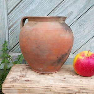 May include: A rustic, terracotta-colored clay pot with a handle, sitting on a wooden surface next to a red apple.