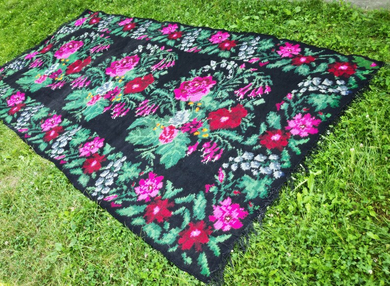 May include: A black rug with a floral pattern featuring pink, red, and green flowers. The rug has a fringed edge.