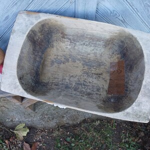 May include: A large, wooden dough bowl with a worn, rustic finish. The bowl is oval-shaped and has a deep well. The wood is a light brown color and has a natural grain pattern. The bowl is sitting on a wooden surface.