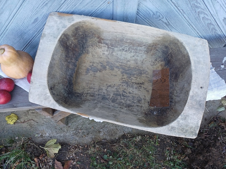 May include: A large, wooden dough bowl with a worn, rustic finish. The bowl is oval-shaped and has a deep well. The wood is a light brown color and has a natural grain pattern. The bowl is sitting on a wooden surface.