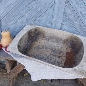 May include: A large, wooden, oval-shaped dough bowl with a rustic, weathered finish. The bowl is empty and sits on a wooden surface with a white cloth draped over it.