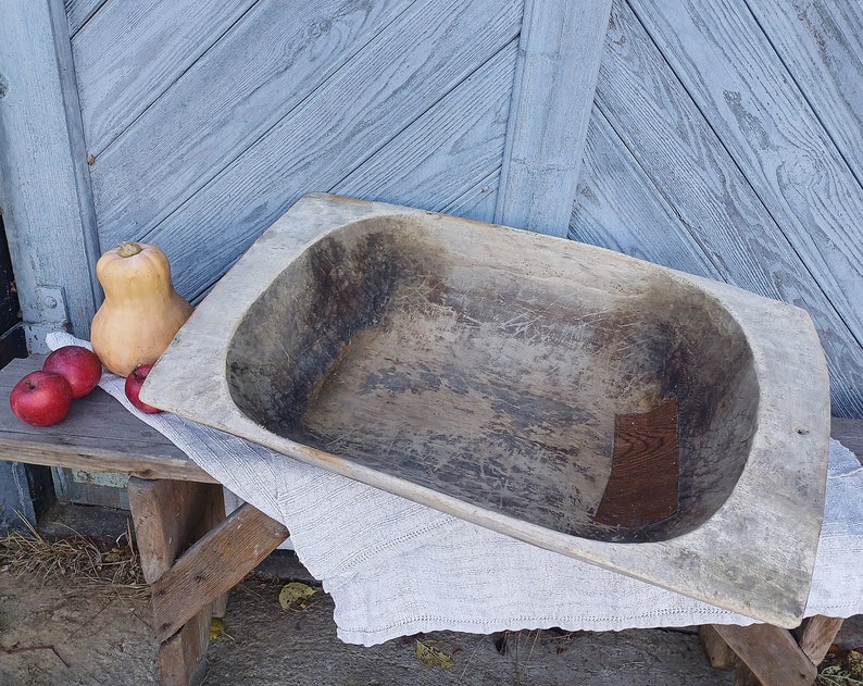May include: A large, wooden, oval-shaped dough bowl with a rustic, weathered finish. The bowl is empty and sits on a wooden surface with a white cloth draped over it.
