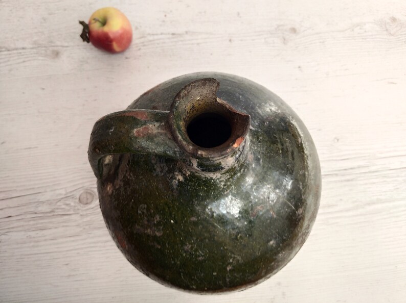 May include: A green glazed ceramic jug with a handle and a chipped opening. The jug has a rustic, aged appearance.