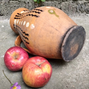 May include: A terracotta-colored ceramic pitcher with a handle, decorated with black and white painted designs. Two red apples rest beside the pitcher. The pitcher has a dark, circular base. The scene is set on a concrete surface.