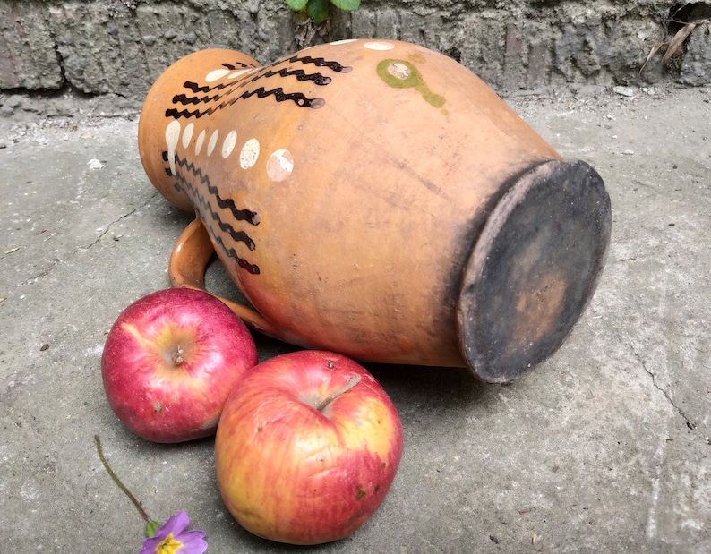 May include: A terracotta-colored ceramic pitcher with a handle, decorated with black and white painted designs. Two red apples rest beside the pitcher. The pitcher has a dark, circular base. The scene is set on a concrete surface.