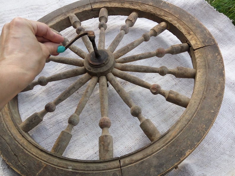 May include: A large, wooden spinning wheel with a circular frame and spokes. The wheel is made of natural wood and has a rustic appearance. The spokes are connected to a central hub, and the wheel is designed for spinning yarn.