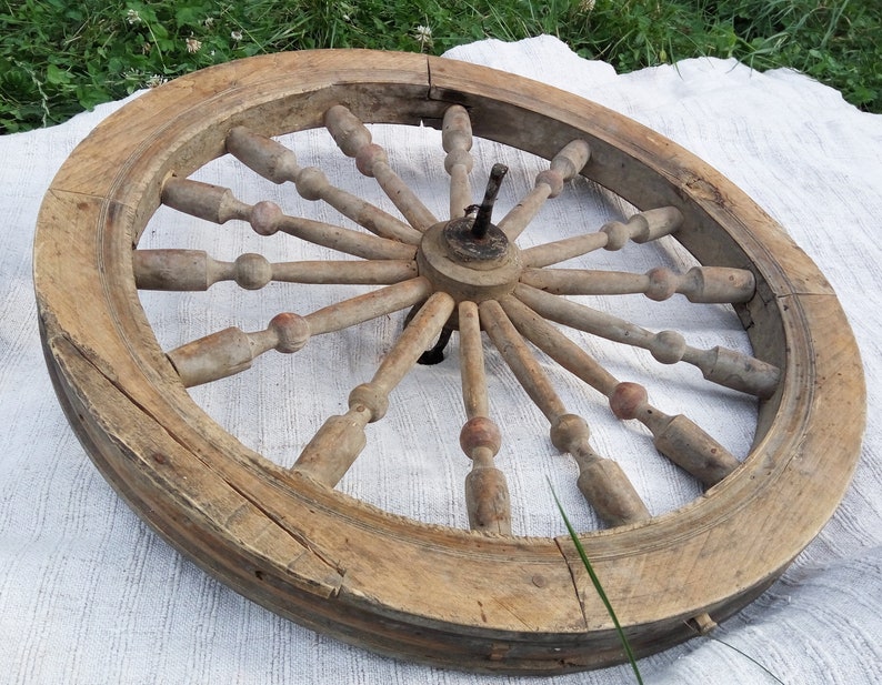 May include: A large, wooden spinning wheel with a circular frame and spokes radiating from the center. The wheel is made of natural wood and has a rustic, antique appearance.