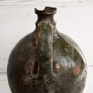 May include: A large, green, ceramic jug with a chipped rim and a handle. The jug has a rustic, aged appearance with a mottled green glaze and white spots.
